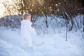 Baby playing with teddy in the snow, winter time. Little toddler boy in handmade white snowsuit, holding teddy bear on sunset, playing outdoors in winter park