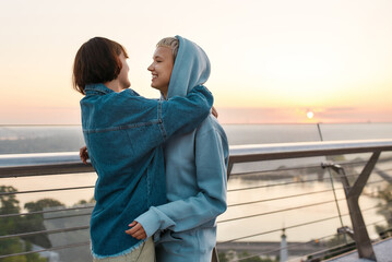 Young loving lesbian couple hugging, looking at each other while enjoying romantic moments together, standing on the bridge at sunrise