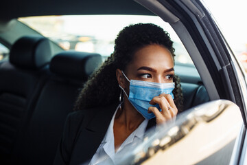 Young businesswoman being a taxi passenger and having a ride wearing and adjusting medical mask for health protection. Business trips during pandemic, new normal and coronavirus travel safety concept.