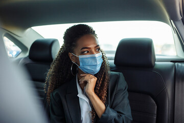Young attractive businesswoman having a ride on a backseat of a taxi during covid-19 pandemic wearing mask. Business trips during pandemic, new normal and coronavirus travel safety concept.