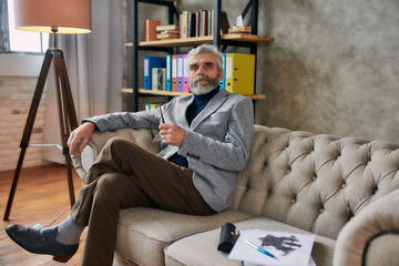 The best counseling. Portrait of mature psychologist looking at camera, holding glasses sitting on...