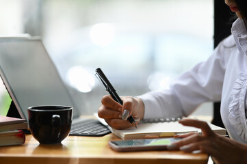 Closeup woman hand working at a smartphone and writing on a notepad with a pen in the office.