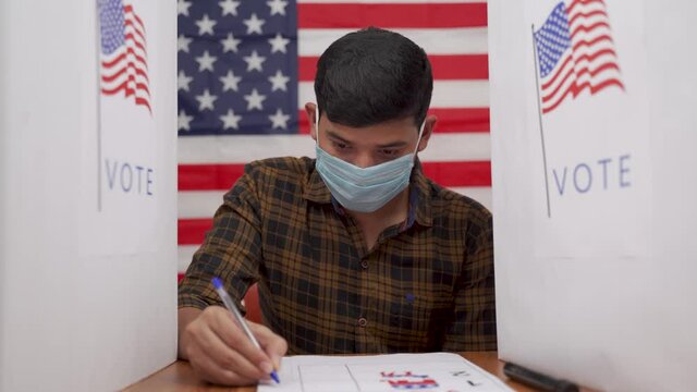 Young man in medical mask busy inside the polling booth with US flag as background - Concept of in person voting with covid-19 safety measure at US election