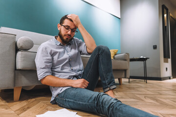 Young confused man analyzing finances at home, holding his head with hand and looking at documents. Sitting on floor near sofa in living room.