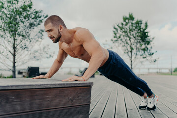 Obraz premium Confident muscled sportsman in sport wear doing push up outdoors, works on biceps and trains determination. Strong topless athletic man exercises in open air. People, motivation, training concept