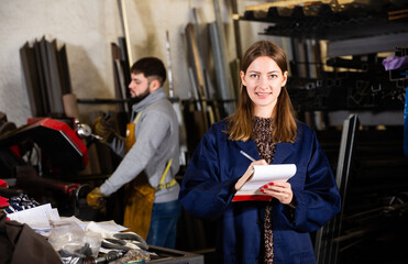 Professional young woman engineer taking notes in notebook in workshop, man on background..