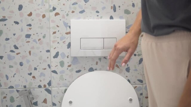 A woman's hand presses a white flush button in a suspended toilet. A woman washes off the toilet at home