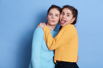 Naklejka premium Two positive Caucasian young girlfriends hugging and having fun while posing isolated over blue background, ladies making grimace and showing tongue, wearing casual attires.