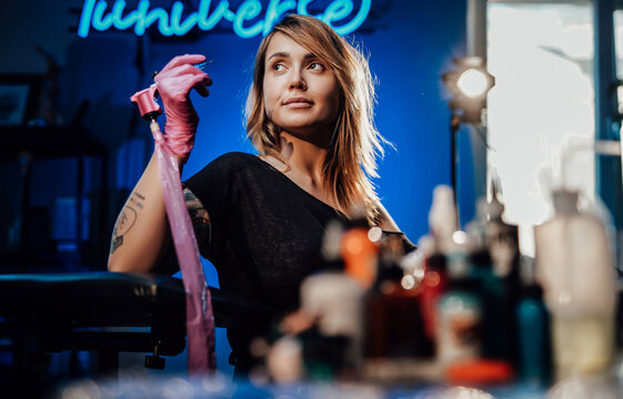 Modern tattoo studio with spotlight and neon lights and attractive female tattoo master shes posing in it with gloves and tattoo device.