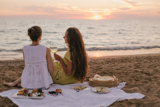 Two young beautiful women female friends having picnic with wine, pizza, cheese and fresh fruits on a beach near ocean.