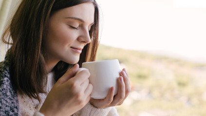 Young woman in white knitted sweater drinking tea near window