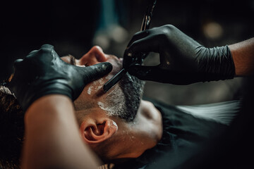 Barber in black gloves maintains hygiene when cutting a beard of his client with sharp razor in...