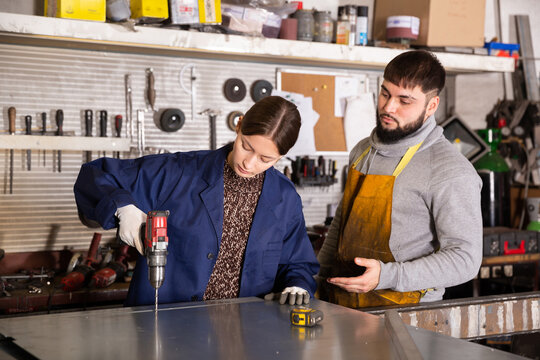 Experienced bearded foreman controlling work of young workwoman drilling holes with hand cordless drill in metal structures in metalworking workshop