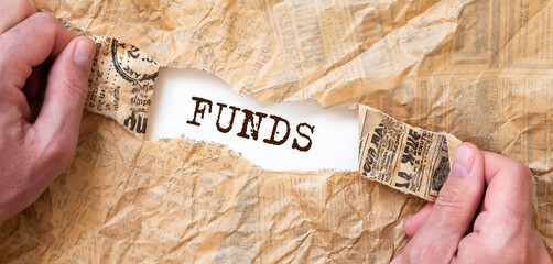 FUNDS word on white table through torn craft brown paper with male hands, top view