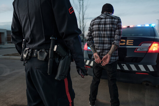 A Police Officer watches a handcuffed suspect at the back of his cruiser
