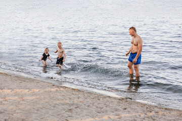 A sports man of European appearance plays with his children in the water. The family bathes in the lake. Father, son and daughter. Holidays and games. Water splash. Lakes in the summer at sunset.