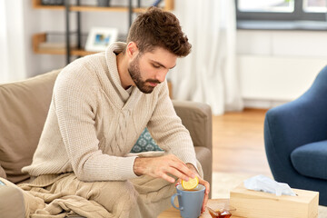 health, cold and people concept - sick young man in blanket drinking hot tea with lemon and honey at home