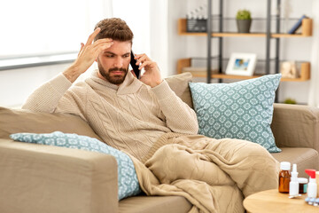 health, cold and people concept - sick young man in blanket with smartphone and medicine calling doctor at home