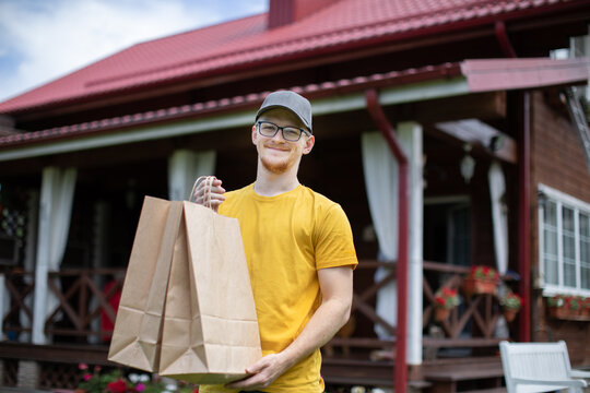 Delivery right to your door. Handsome smiling young delivery man in glasses holds paper bags of food in open air Looking at camera. Rustik style red roof country cottage on blurred background.