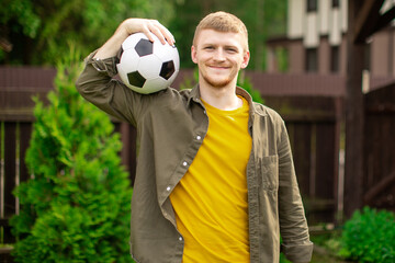 young caucasian man in everyday clothes carry soccer ball on his shoulder on green lawn of country house. amateur football invitation