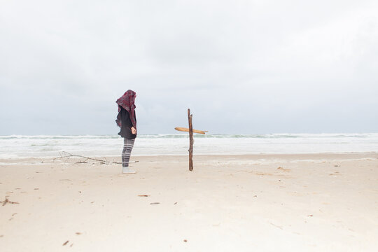 Sad woman standing next to a wooden cross on the beach.