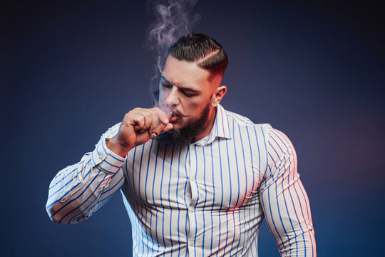 Handsome and elegance bodybuilder with beard and fashionable hairstyle smoking cigar in studio background.