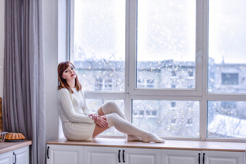 Obraz na płótnie Canvas A beautiful young woman in a white knitted sweater dress and long leggings, sits on a wide windowsill at the window. Looking into the distance, sad, loneliness concept. Copy space. White interiors