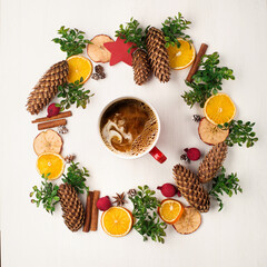 Cup of hot coffee with milk, green branches, pine cones, oranges and apples on white wooden background.