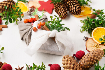 Stylish rustic gifts wrapped in linen fabric with green branches on wooden table with pine cones, oranges and apples.
