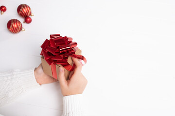 Christmas holiday shopping concept. Small craft gift box with red ribbon in girl's hands. White background, Several Christmas balls are visible. Place for your text. Horizontal. 