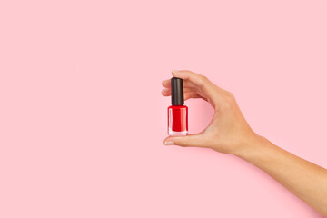 Mano femenina sosteniendo una botella de esmalte de uñas  de color rojo sobre un fondo rosa pastel liso y aislado. Vista de frente. Copy space. Concepto: Belleza.