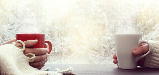 red and white mugs in hands against the background of a frosty window. a warming drink for a winter...