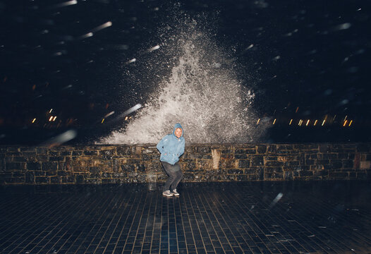 Man crouching down to shelter from the water splashing up against the harbour walls