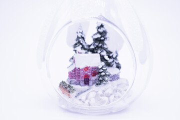 Close up of the small house in a snowy forest among the firs inside the transparent Christmas ball, White background, copy space. Cozy winter holiday conception