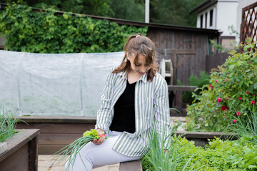 Young beautiful woman gardener sitting on raised garden bed harvests fresh herbs green leek and lettuce, farmer working in garden in countryside. Agriculture, farming, organic products, food growing