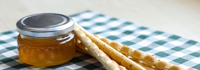 Breadsticks or Italian grissini snack with jar of honey on green gingham tablecloth. Food, harvest concept. Banner.