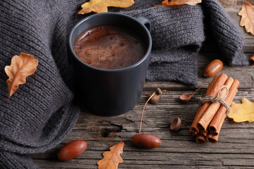 Black coffee mug, yellow oak leaves, acorns, tied cinnamon sticks, grey scarf on rough old wooden table. Closeup, high angle. Autumn, fall season, thanksgiving, coffee shop menu, hot drink concept