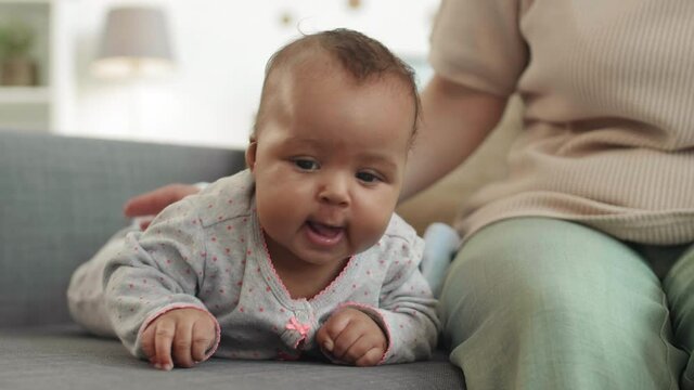 Close up of mixed-race cute toddler is lying on couch at home near unrecognizable woman sitting nearby