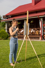 attractive girl paints easel painting at her country house outdoor. Pretty woman is painting. Open air session. Outdoor time for hobbies