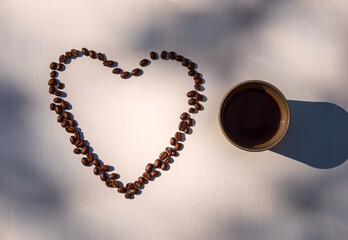 Open heart formed with coffee beans next to a cup of coffee lit by evening light causing soft shadows