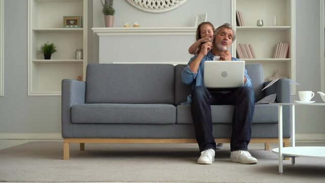 Busy caucasian father using laptop for distant work with daughter in living room. Upset child wants to play with dad