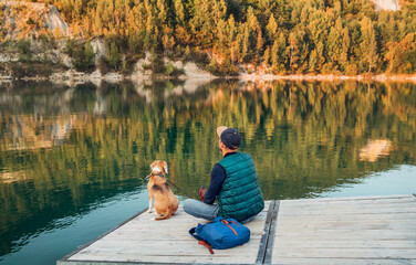 Man as a dog owner and his friend beagle dog are sitting on the wooden pier on the mountain lake and enjoying the landscape during their walking in the autumn season time. Human and pet concept image.