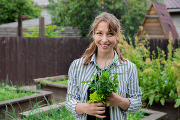 Young beautiful farmer with fresh harvest herbs in hands, happy woman gardener standing on garden background looks at camera with smile, Home organic eco products - green leek, lettuce, parsley, dill