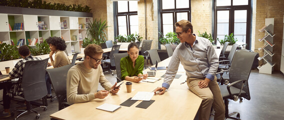 Sharing fresh ideas. Three young colleagues sitting at the desk in the modern coworking space, chatting and discussing new project, business people working together - obrazy, fototapety, plakaty