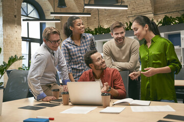 Group of young happy multicultural business people looking at laptop screen and discussing work or...