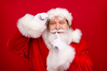 Photo of retired old man grey beard take off eyeglasses finger cover lips excited want surprise kid wear x-mas costume white gloves coat spectacles headwear isolated red color background
