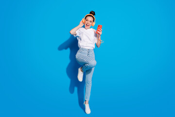 Full length photo of cheerful crazy girl jump use smartphone listen sound melody headset look...