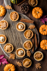 Top down view of various pumpkin pie tarts on a wooden platter, surrounded by autumn decorations.