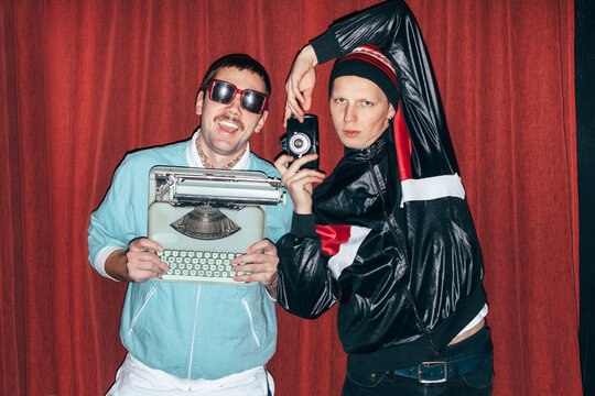 Two young crazy funny men posing on red curtain background with typewriter and old fim camera