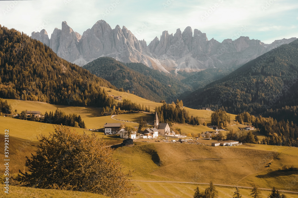 Poster aerial shot of a village on a dry grassy hill surrounded by the forested mountains at daytime